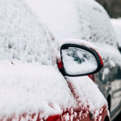 Hóval vastagon borított, utcán parkoló autók téli környezetben, lefagyott visszapillantó tükörrel az előtérben.
