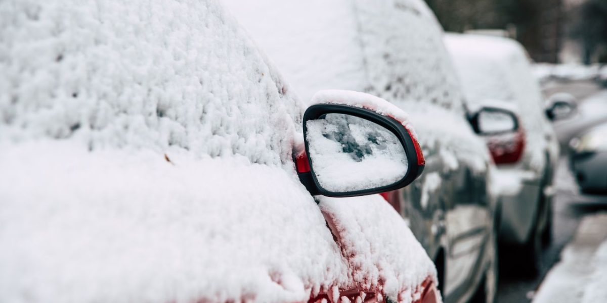 Hóval vastagon borított, utcán parkoló autók téli környezetben, lefagyott visszapillantó tükörrel az előtérben.