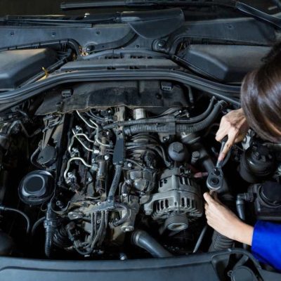 Szerelő nyitott motorháztetőnél dolgozik, az autó motorterében alkatrészeket ellenőriz szerszámmal