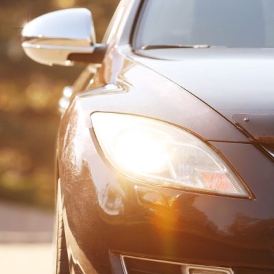 Elölről fotózott személyautó bekapcsolt fényszóróval, őszi környezetben, napfényben haladva az úton.