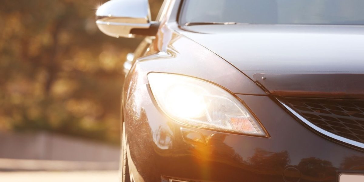 Elölről fotózott személyautó bekapcsolt fényszóróval, őszi környezetben, napfényben haladva az úton.